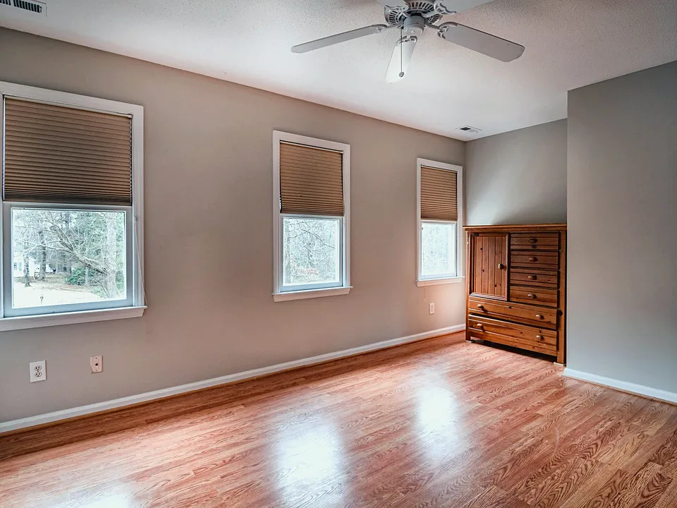 Empty bedroom with three windows, a light wood-look floor and a tall wooden armoire against the wall