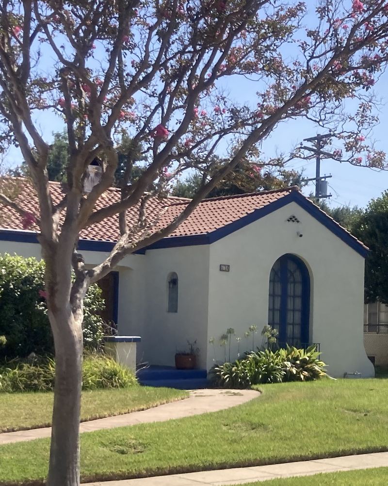 White stucco Spanish-style home in Alhambra with a red clay tile roof, blue trim and an arched front window, behind a flowering crape myrtle